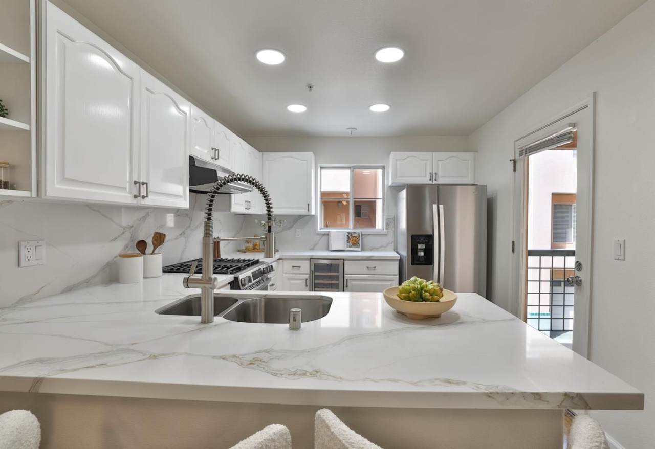 Second view of the kitchen at counter height, marble-veined quartz sweeping to a stainless-steel refrigerator and a small side balcony beyond