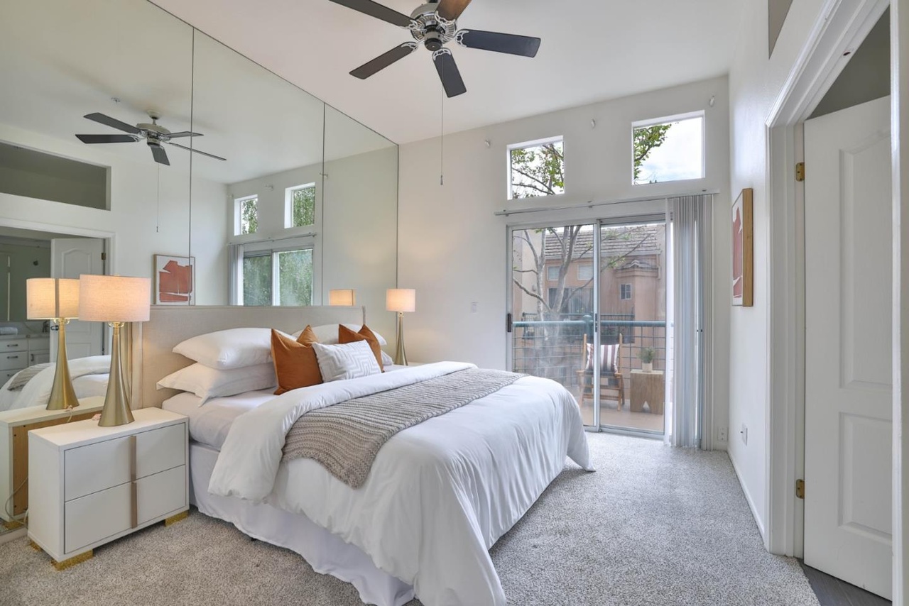 Primary bedroom with a vaulted ceiling, a full-wall mirror behind the bed, a king bed dressed in white, and sliding glass doors to a private balcony with a chair set out