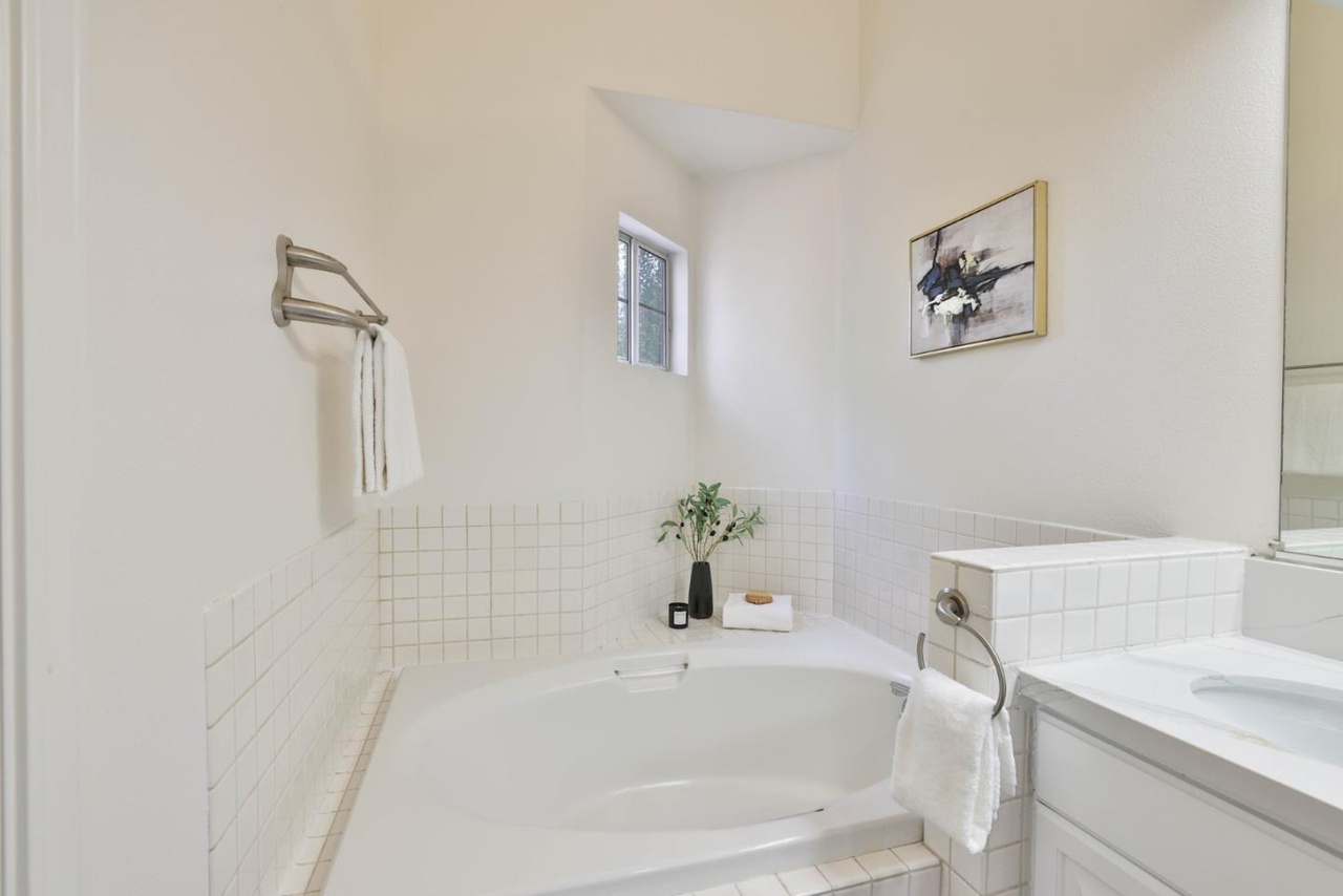 Primary bath with an oversized corner soaking tub set in white tile, a small awning window above, and a framed print on the side wall